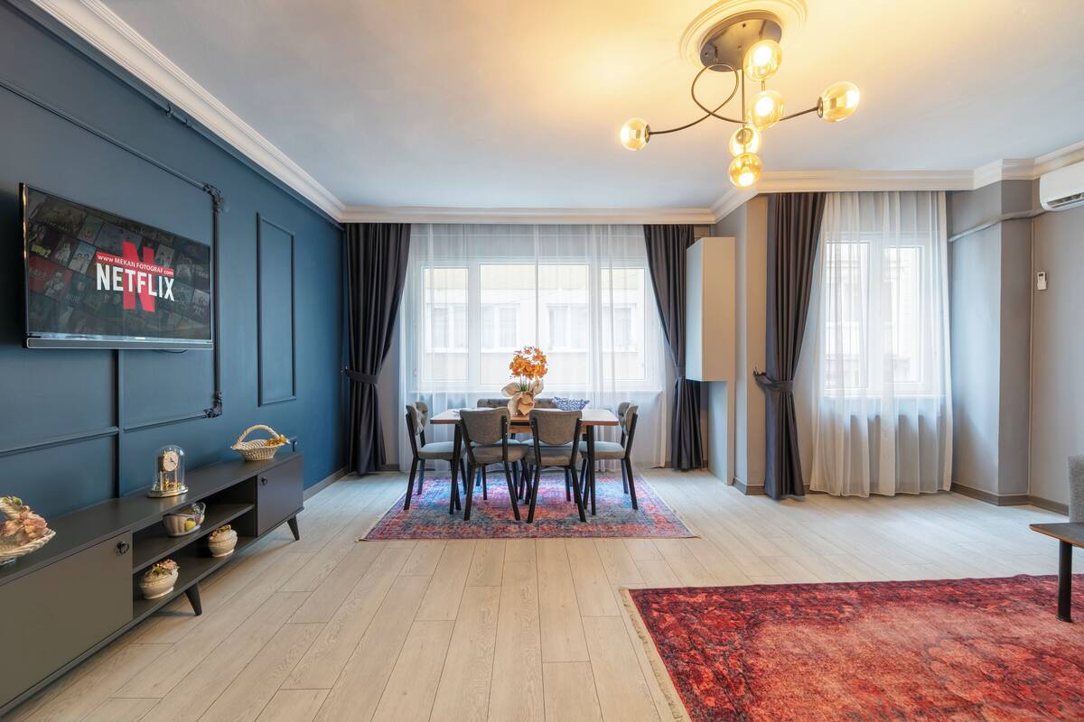 A modern living room with a dining area, a flat-screen TV mounted on the wall, a chandelier, and a red rug.