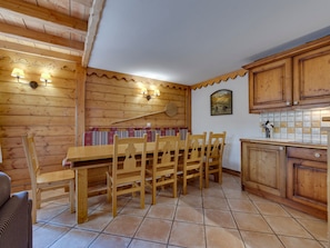 A wooden dining area with a long table and chairs, a bench with cushions, and a tiled kitchen area with wooden cabinets.