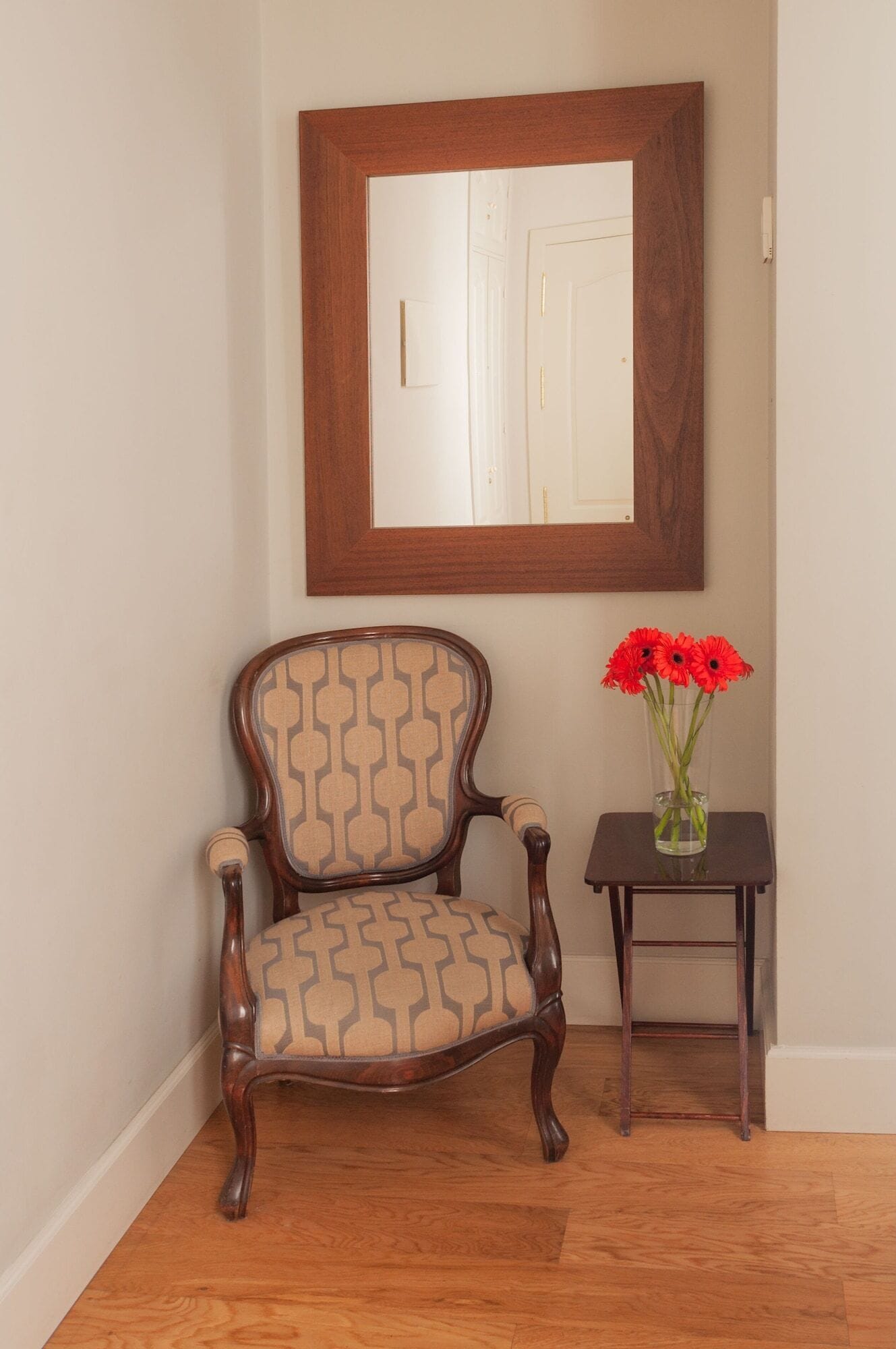 A wooden chair with a patterned cushion, a small table with a vase of red flowers, and a mirror on the wall.