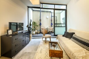 A modern living room with a sofa, a coffee table, a TV, and a cabinet. There is a balcony with plants and a view of the outside. - 1BR duplex Apt with private Balcony (Tel Aviv)