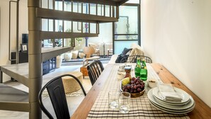 Une salle à manger avec une nappe à carreaux, des assiettes, des verres et un saladier de raisins. On aperçoit un escalier au fond, qui mène à un autre niveau.