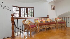 Un escalier avec une rampe et des balustrades en bois, menant à une pièce où se trouvent deux canapés en osier ornés de coussins à motifs, une grande fenêtre avec des vitraux et un motif mural représentant des oiseaux.