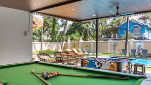 Une table de billard, une table de baby-foot, des queues de billard et des billes, le tout installé dans un espace abrité avec vue sur une piscine et des chaises longues.
