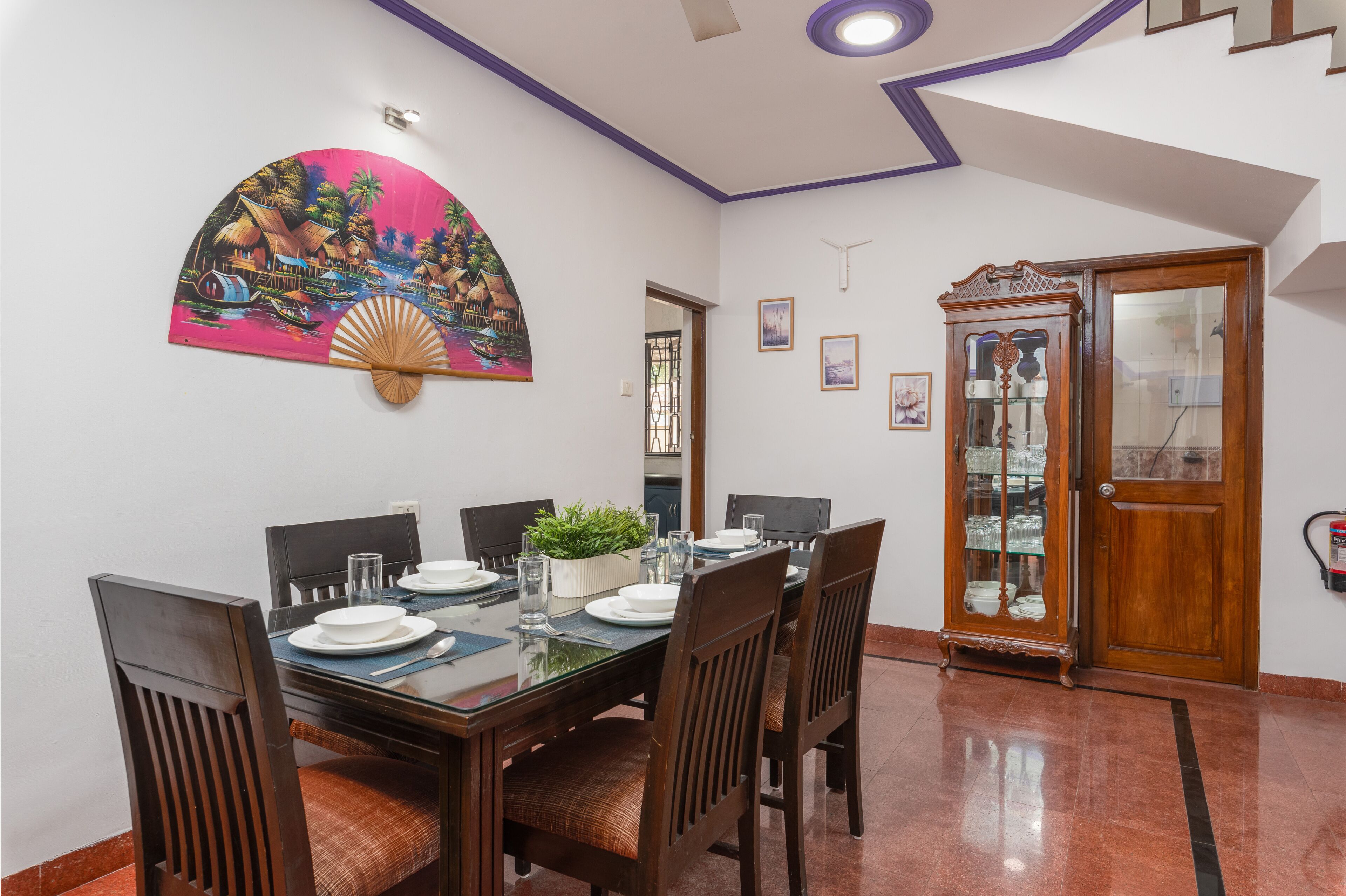 Un comedor con una mesa de madera dispuesta para cuatro personas, rodeada de sillas. Un abanico decorativo cuelga de la pared, y se ve un aparador con puertas de cristal al fondo.