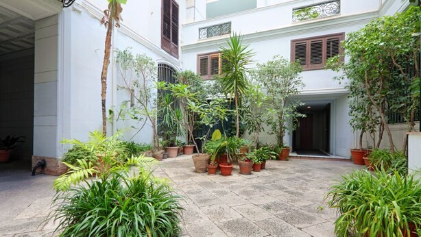 A courtyard with potted plants, a stone floor, and a building with multiple windows and shutters.