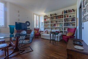 A living room with a glass table, chairs, a sofa, a bookshelf, and a television.