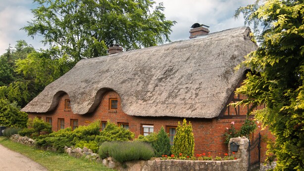 A traditional thatched-roof house with brick walls, surrounded by well-maintained gardens and trees. - Idyllically situated vacation apartment 10 min. to the natural beach (Barkelsby)