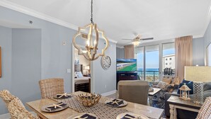 A dining area with a wooden table set for four, surrounded by wicker chairs. A chandelier hangs above, and a television is visible in the background. - Azure 422 ~ Ocean Views ~ 2 Sets of Free Beach Service (Fort Walton Beach)