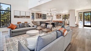 A spacious living area with a grey sofa, a coffee table, and a patterned rug. A kitchen with wooden cabinets and a dining area with chairs is visible in the background. - Pet Friendly | Minutes to Lift | Hot Tub | Blue Mountain Escape (Angel Fire)