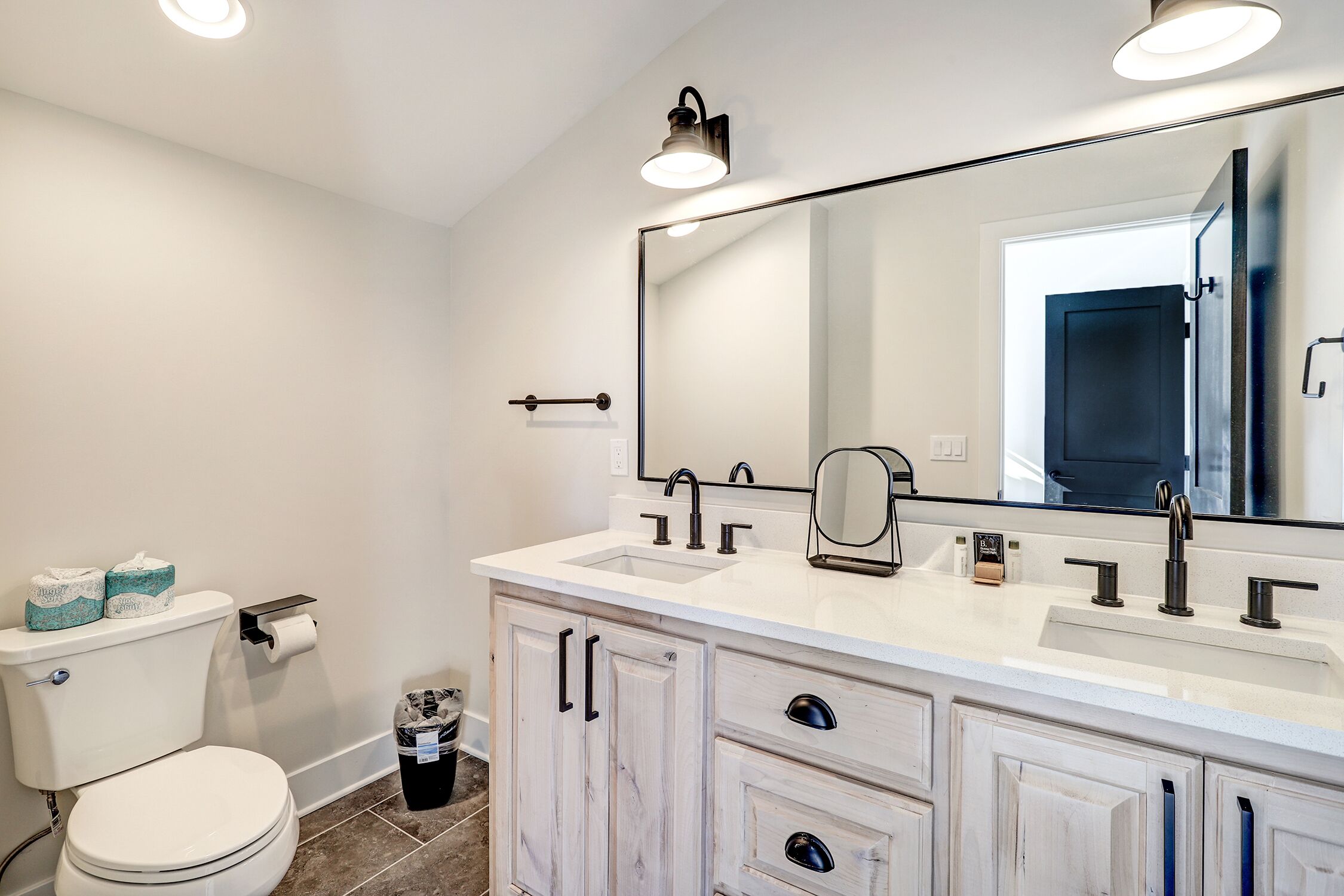 Une salle de bain avec des toilettes blanches, deux lavabos, un grand miroir et des meubles blancs.
