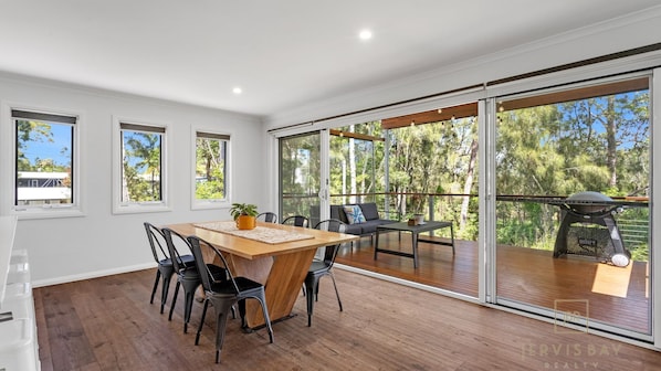 A spacious room with a wooden dining table, black chairs, and a grey sofa. There are large windows and a sliding glass door leading to an outdoor deck with a barbecue grill. - Kookaburra Rest - Vincentia (Vincentia)