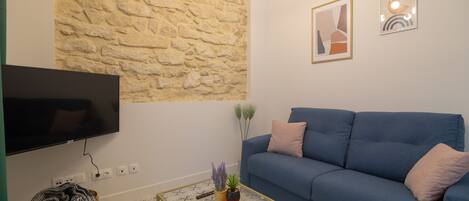 Una sala de estar con una pared de piedra, un sofá azul, una mesa de centro de vidrio con libros, un televisor de pantalla plana y una cesta de mimbre.