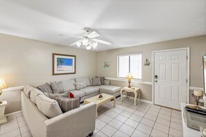 Un salon avec un canapé beige, une petite table, un ventilateur de plafond et une porte.