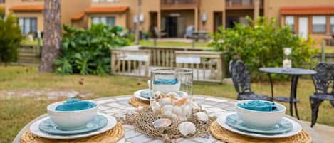 A table set with bowls and shells, a glass, and a woven placemat, with a view of a building with a red roof and balconies.