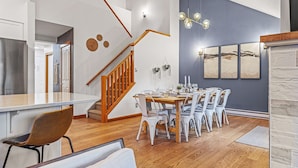 A modern kitchen with a wooden staircase, a dining area with a wooden table and white chairs, and a gray accent wall with framed artwork.