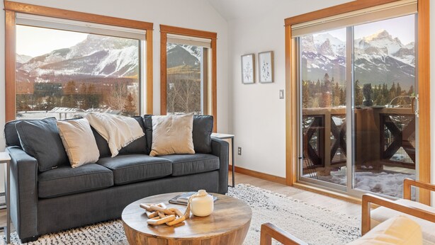 A modern living room with a gray sofa, a round wooden coffee table, and a large sliding glass door leading to a balcony with a mountain view.