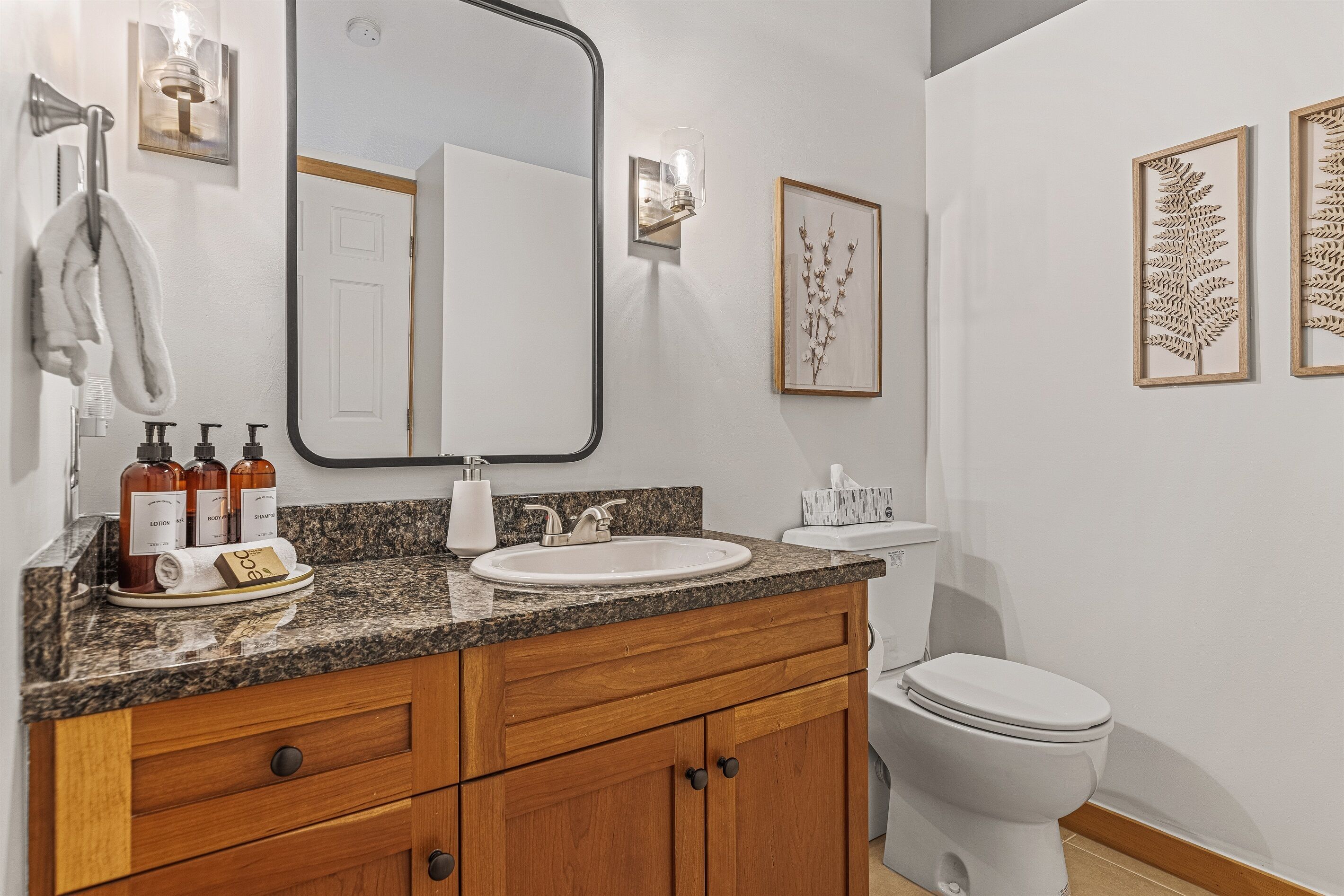 Un baño con encimera de granito, un lavabo blanco, tocador de madera con cajones, inodoro y arte botánico enmarcados en la pared.
