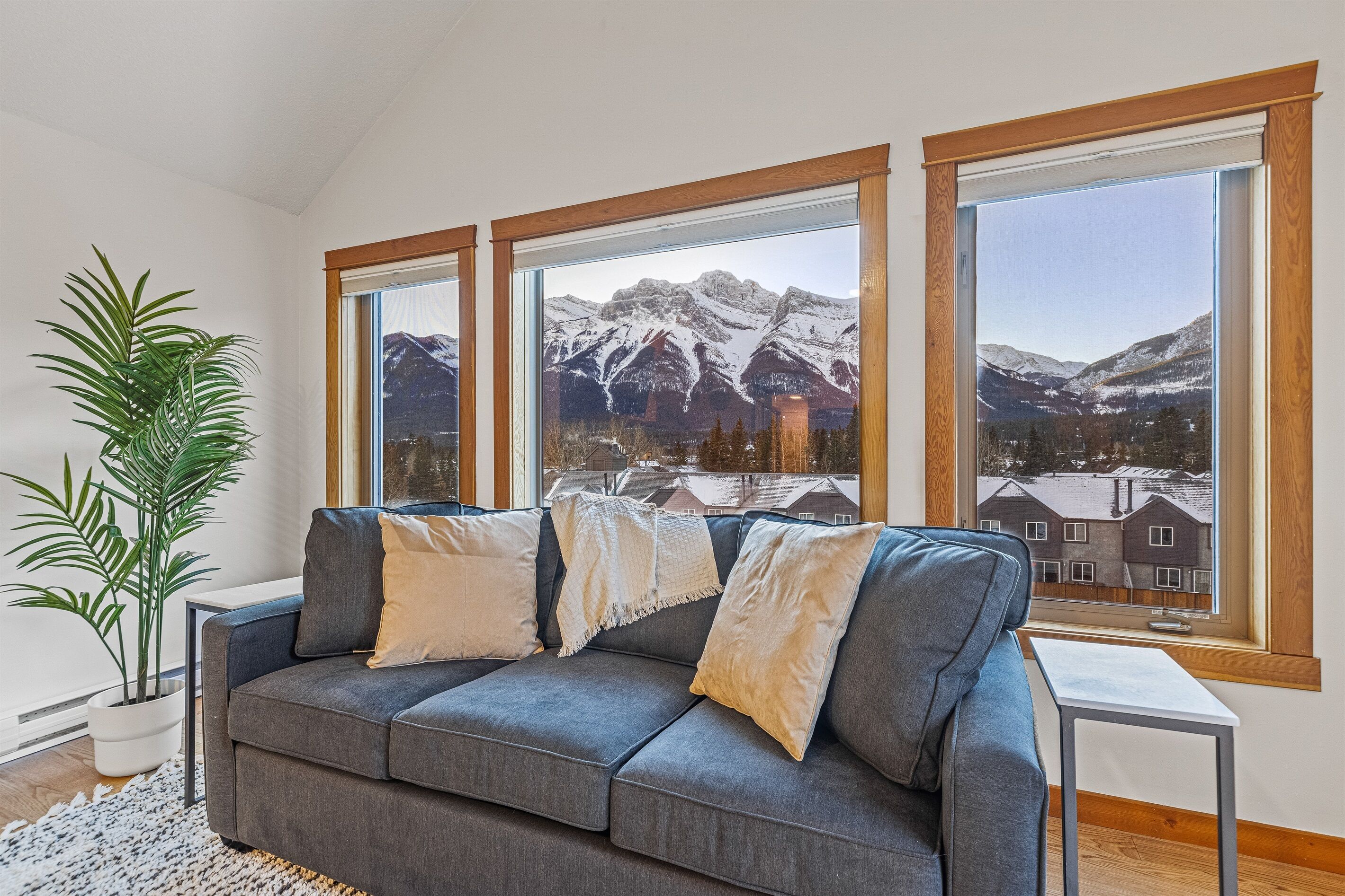 Una sala moderna con un sofá gris, almohadones blancos y un ventanal con vistas a montañas nevadas.