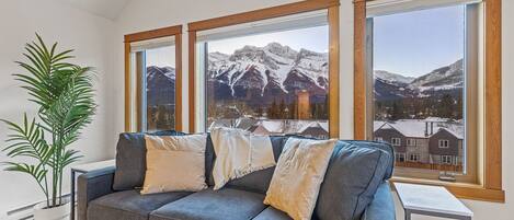 Una sala moderna con un sofá gris, almohadones blancos y un ventanal con vistas a montañas nevadas.