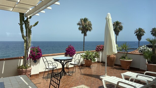 Una terraza con vista al mar, que incluye una mesa, sillas, plantas en macetas y una pérgola.