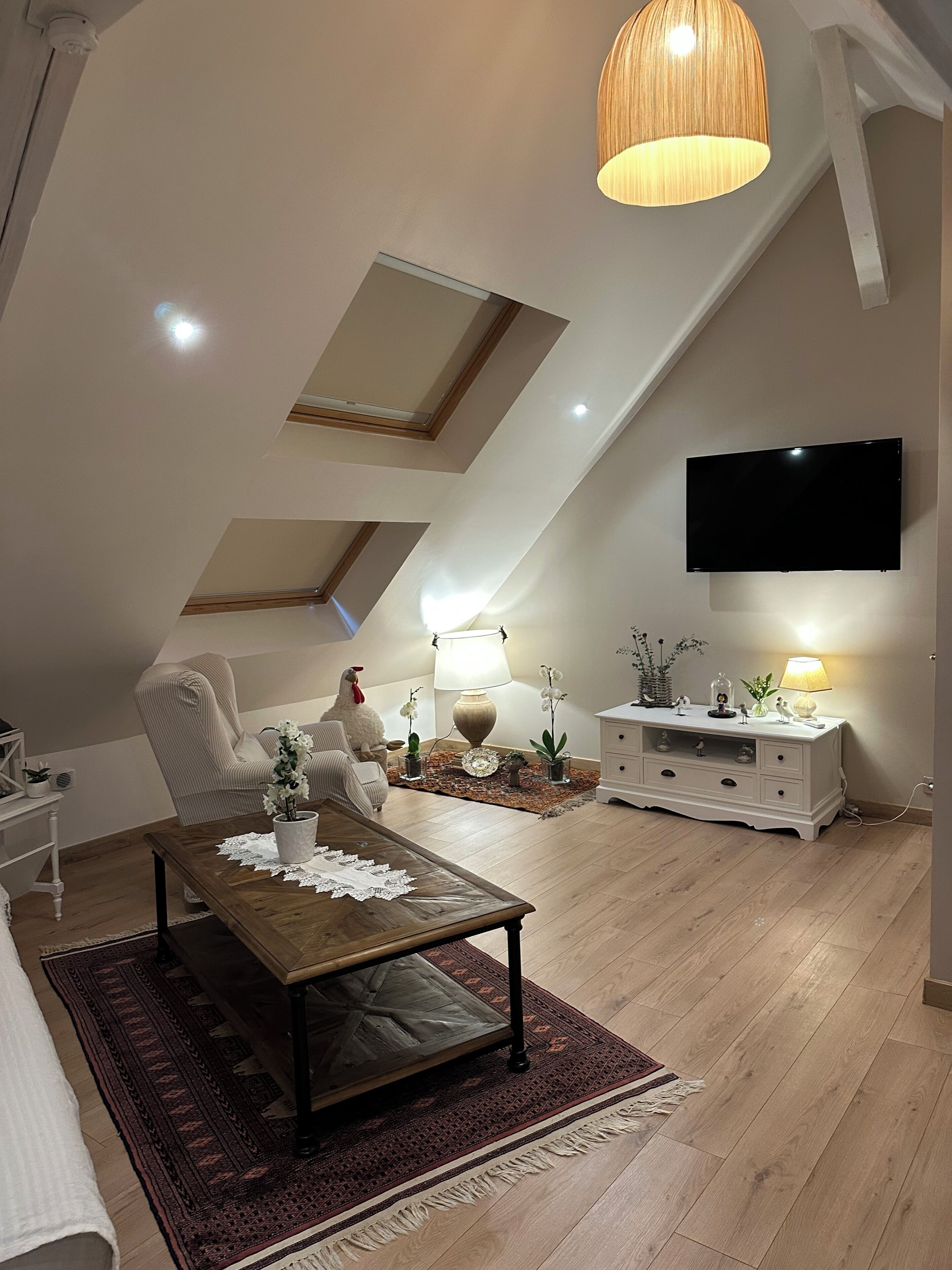 Una habitación tipo loft con techo inclinado, un televisor de pantalla plana en la pared, una mesa de centro de madera, un sofá blanco y una lámpara decorativa.