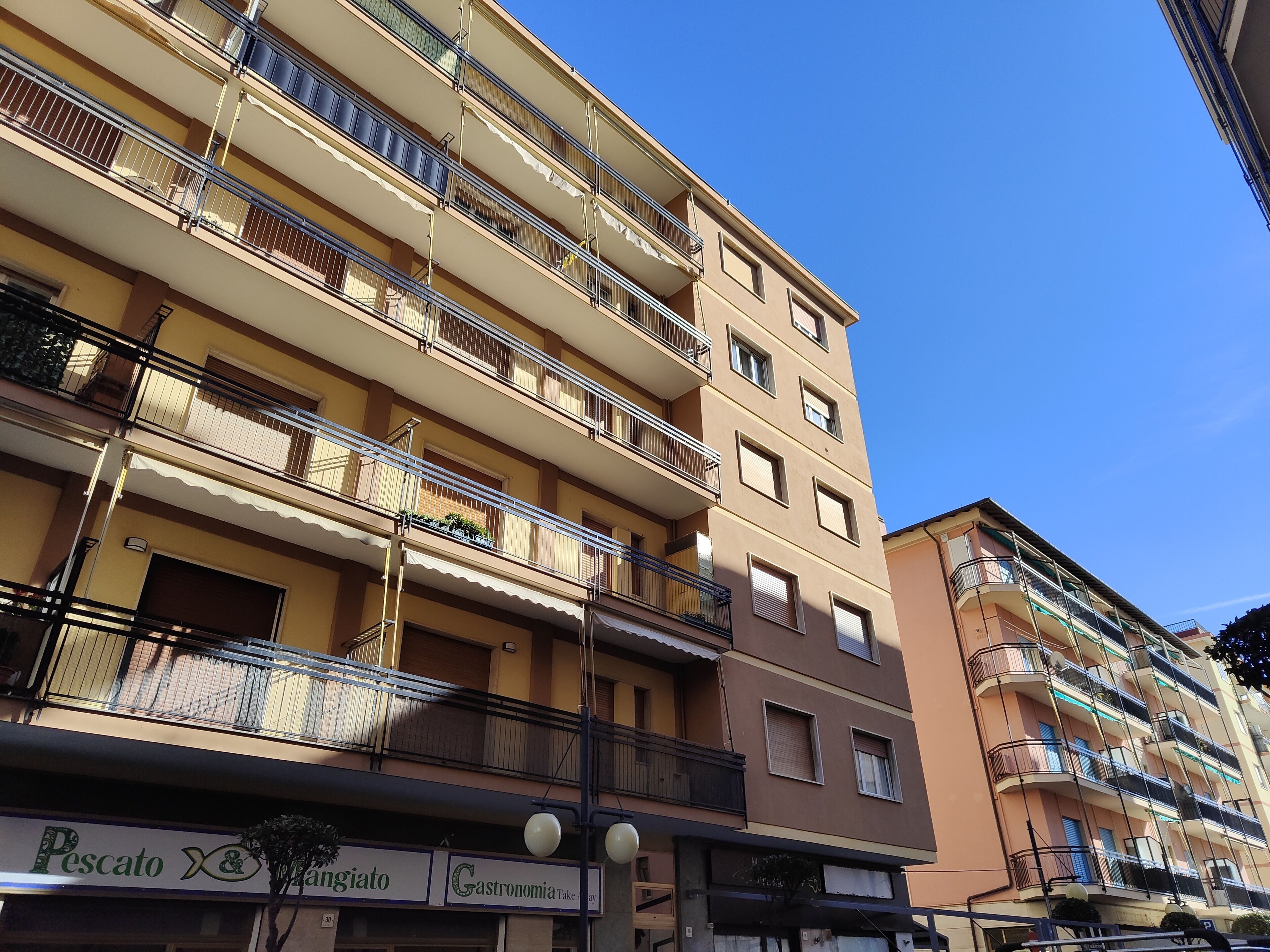 Un edificio de varios pisos con balcones, una tienda con rótulos y un cielo azul despejado.