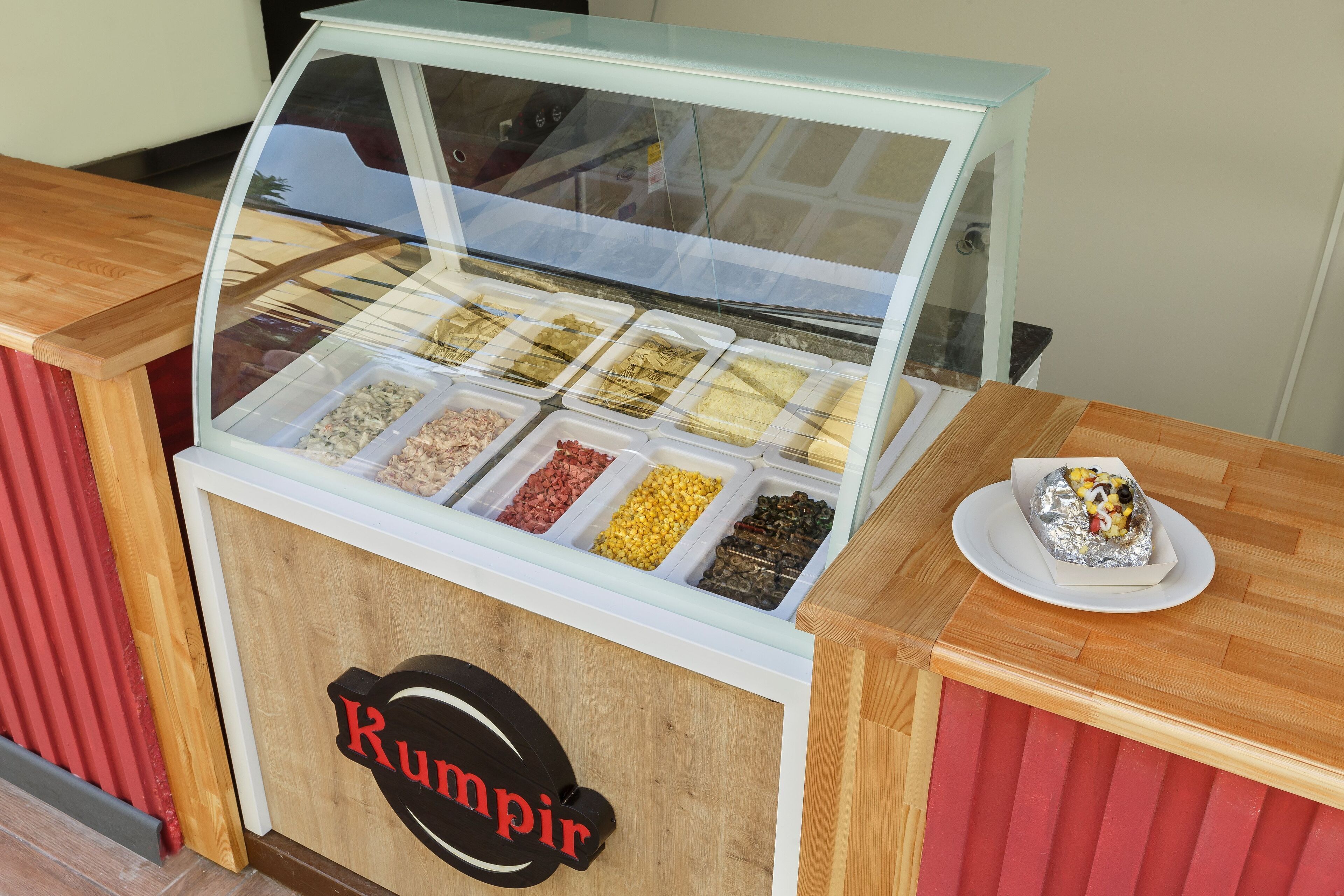 A food display counter with various food items in transparent containers and a sign that reads "Rumpir".