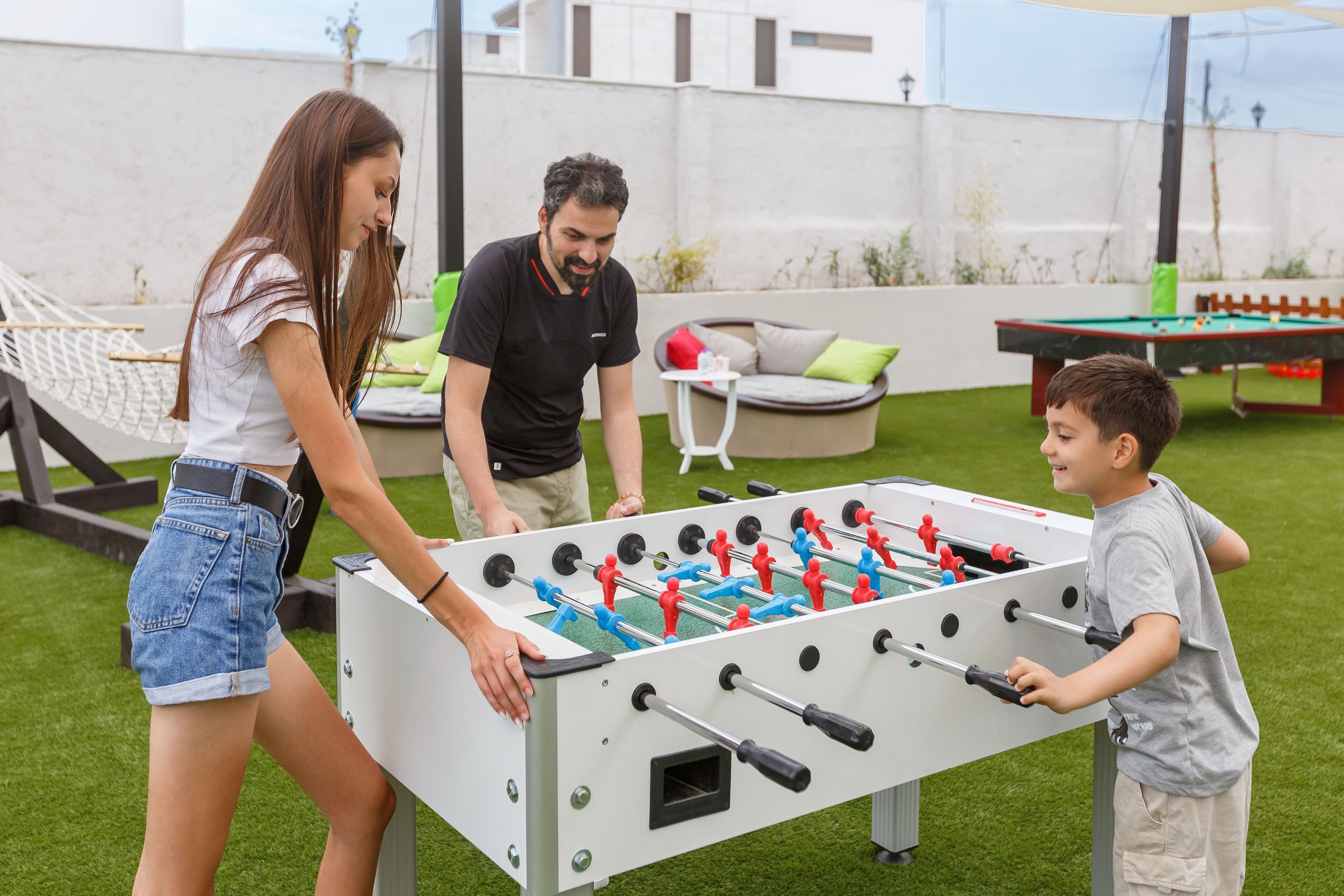 A foosball table in an outdoor setting with a grassy area, a hammock, and a pool table in the background.