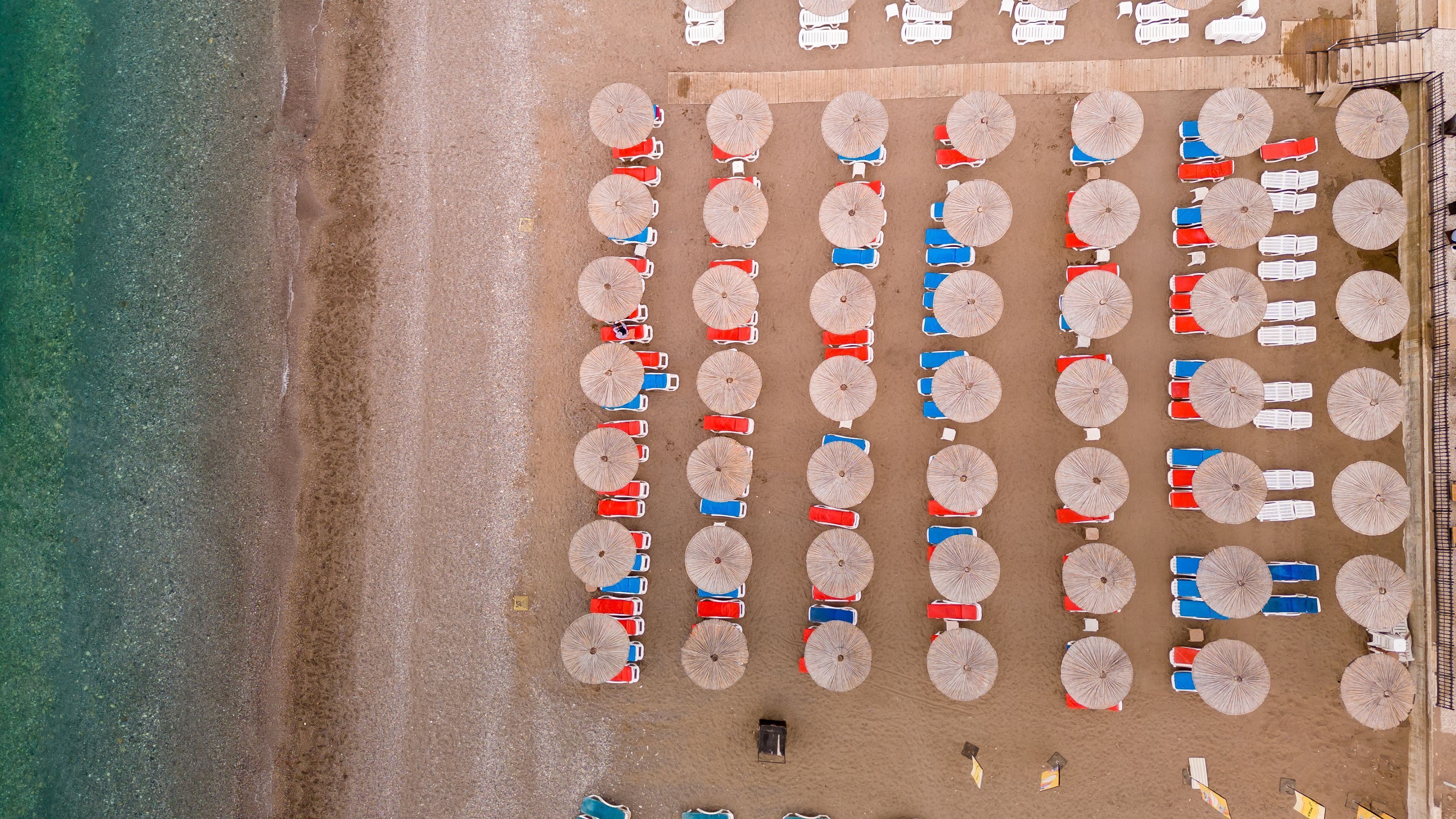 An aerial view of a beach with rows of umbrellas and lounge chairs, a sandy beach, and a clear blue ocean.