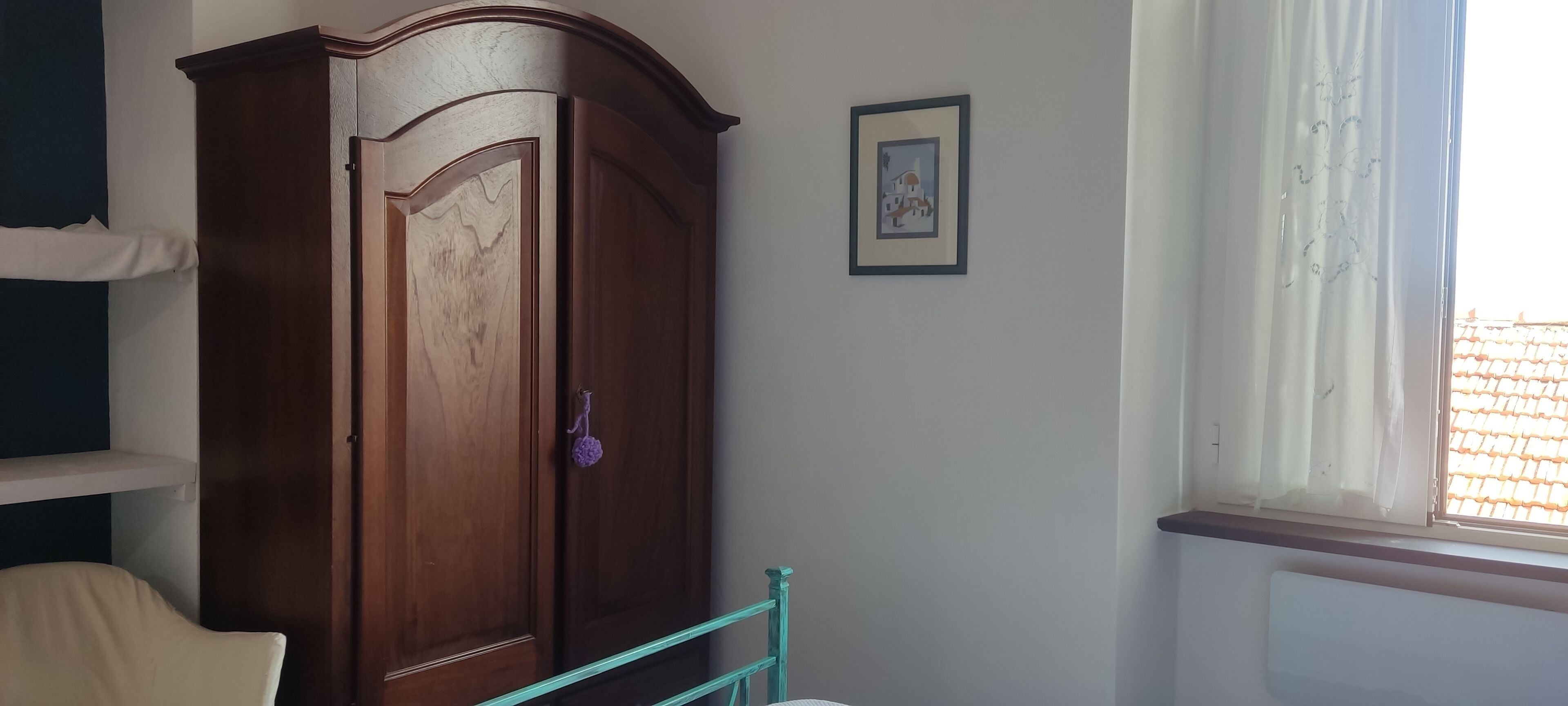 A room with a wooden wardrobe, a framed picture on the wall, and a window with curtains.