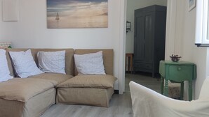 Een knus vertrek met een beige bank, een klein groen bijzettafeltje en een schilderij van een zeilboot aan de muur.