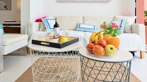 Ein modernes Wohnzimmer mit einem weißen Sofa, einem Couchtisch mit Früchten und einer Flasche, sowie einem Gemälde an der Wand.