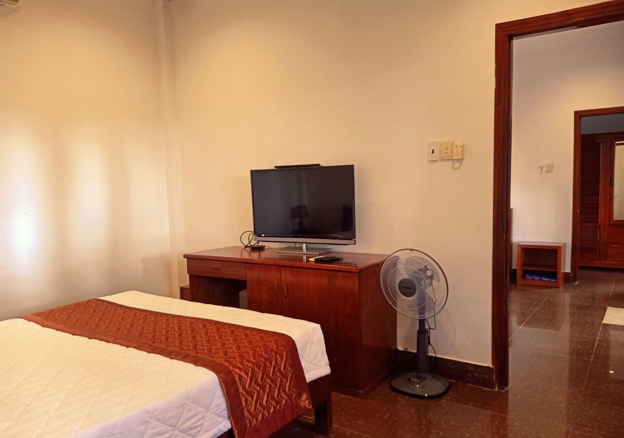 Habitación de hotel con una cama, un televisor sobre un aparador de madera, un ventilador de pie y una puerta que da a otra habitación.
