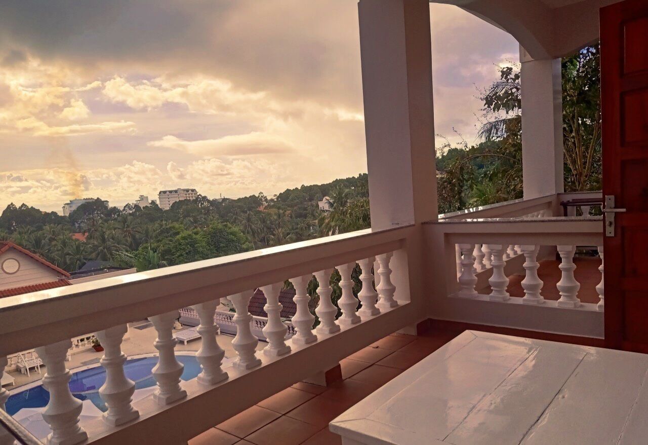 A balcony with a white balustrade, a white table, and a view of a swimming pool and buildings in the distance.