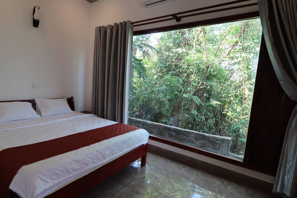 A bedroom with a large bed, a window with curtains, and a view of greenery outside.