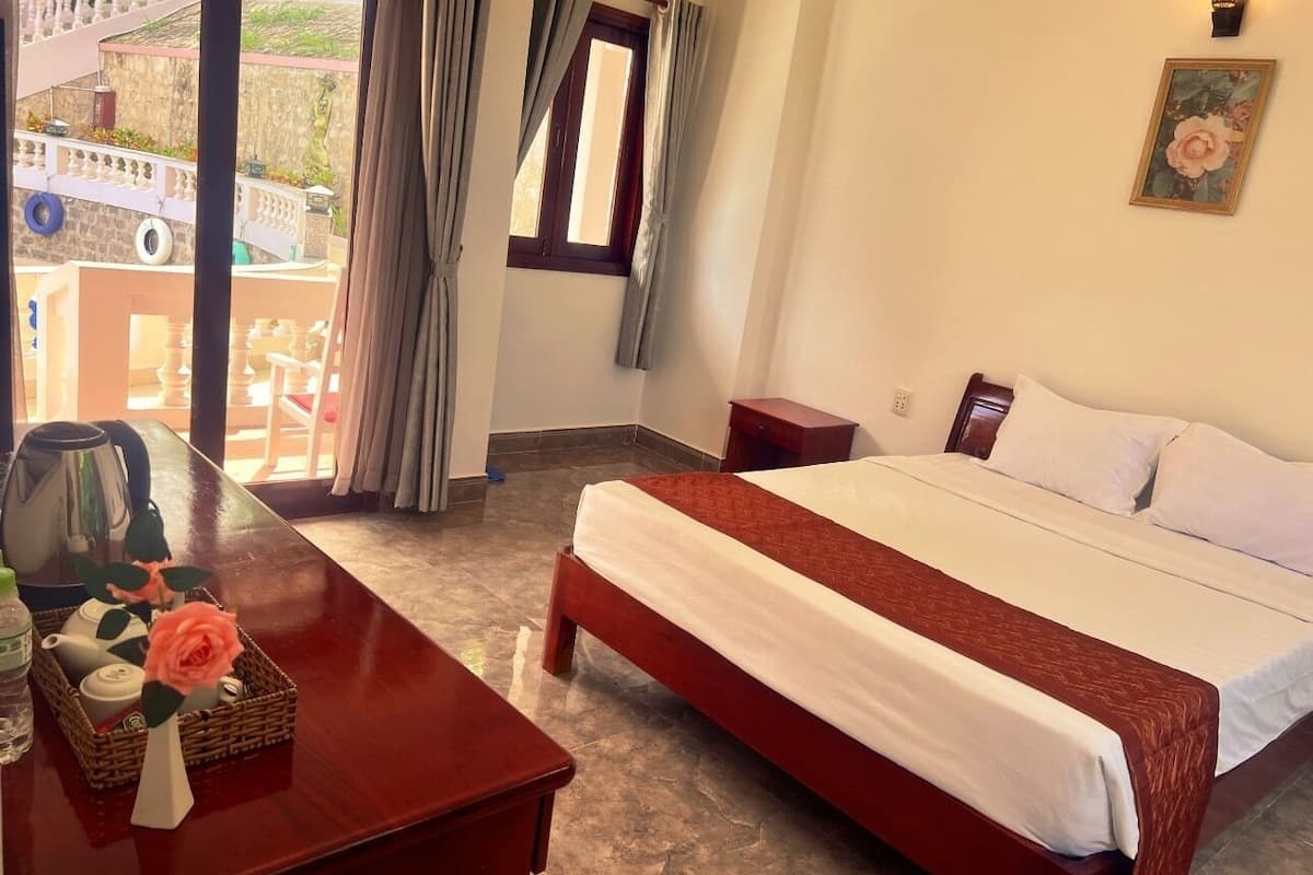 A hotel room with a bed, a desk with a kettle, a vase with a rose, and a view of the outside balcony.