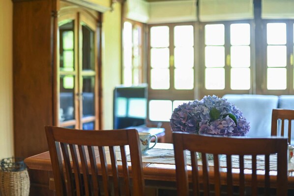 A dining area with a table set for two, a vase of flowers, and a view of a bay window with multiple panes.
