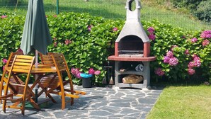 A stone patio with a wooden table and chairs, a stone barbecue grill, and a green umbrella. - Apartamento B (Picones)
