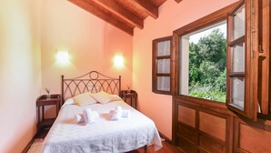A bedroom with a wooden ceiling, a large window with multiple panes, a bed with a white knitted bedspread, two bedside tables with lamps, and a view of greenery outside. - Apartamento B (Picones)