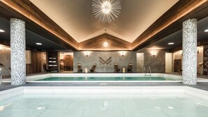 A modern indoor swimming pool with a central seating area, wooden beams, and a unique ceiling light fixture.