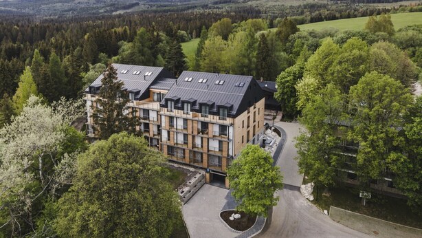 A modern hotel with multiple floors and a flat roof, surrounded by lush greenery and mountains in the background.