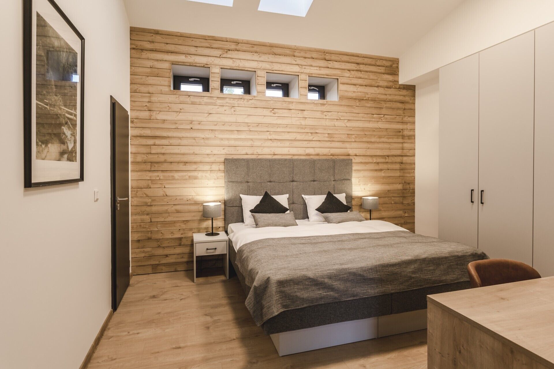 A bedroom with a wooden wall, a bed with a grey headboard, bedside tables with lamps, and a wardrobe.
