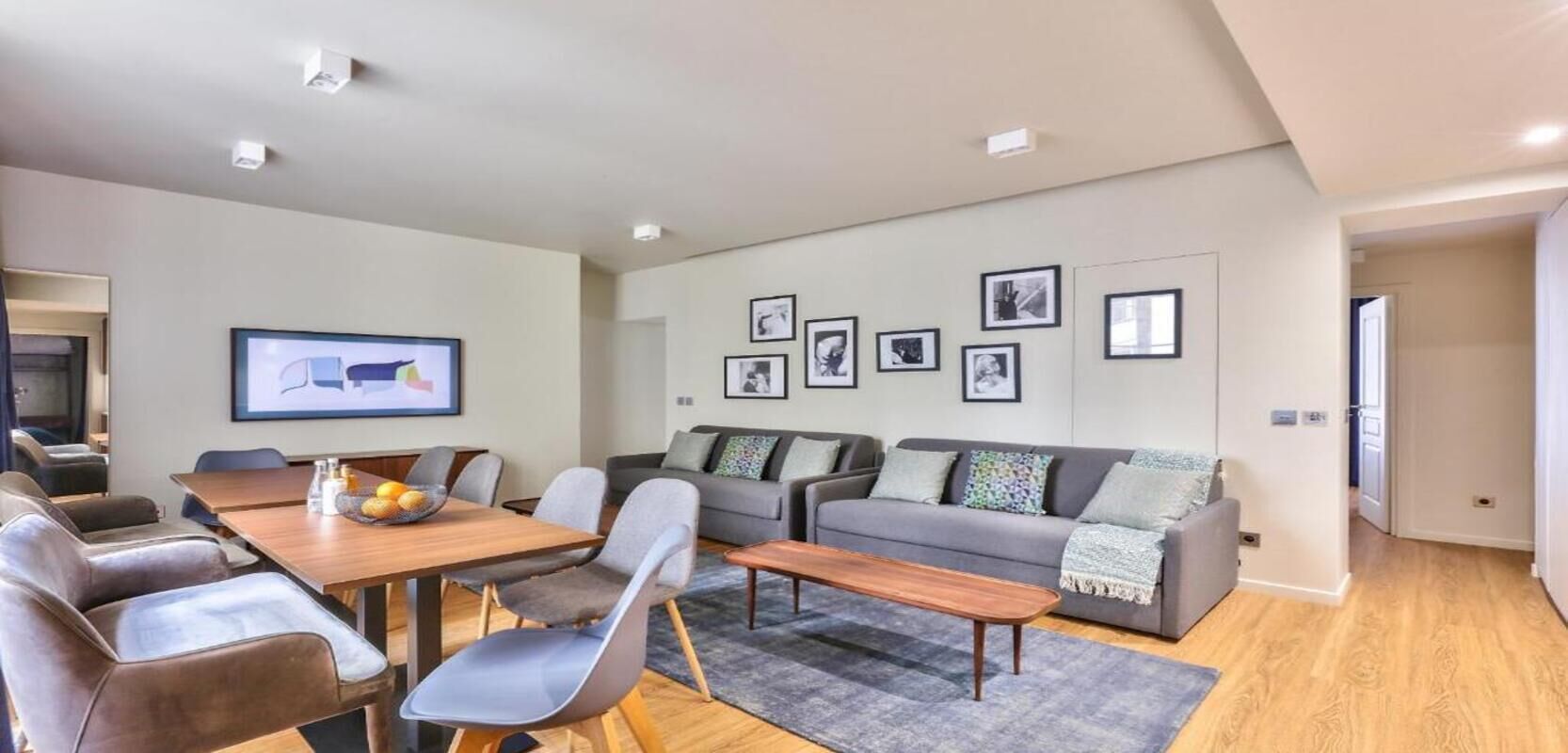 A modern living room with a dining area, grey sofas, a coffee table, and a TV mounted on the wall.