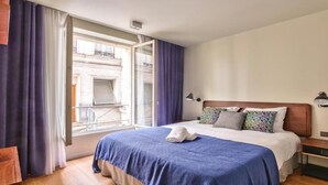 A hotel room with a large bed, a window with curtains, a nightstand, and a view of a building outside. - Elegant Haussmannian Retreat in the Heart of Paris (Paris)