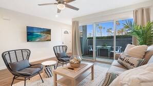 Ein Wohnzimmer mit Deckenventilator, großem Fernseher, einer Couch mit Strandkissen und Blick auf Palmen durch eine Glastür.