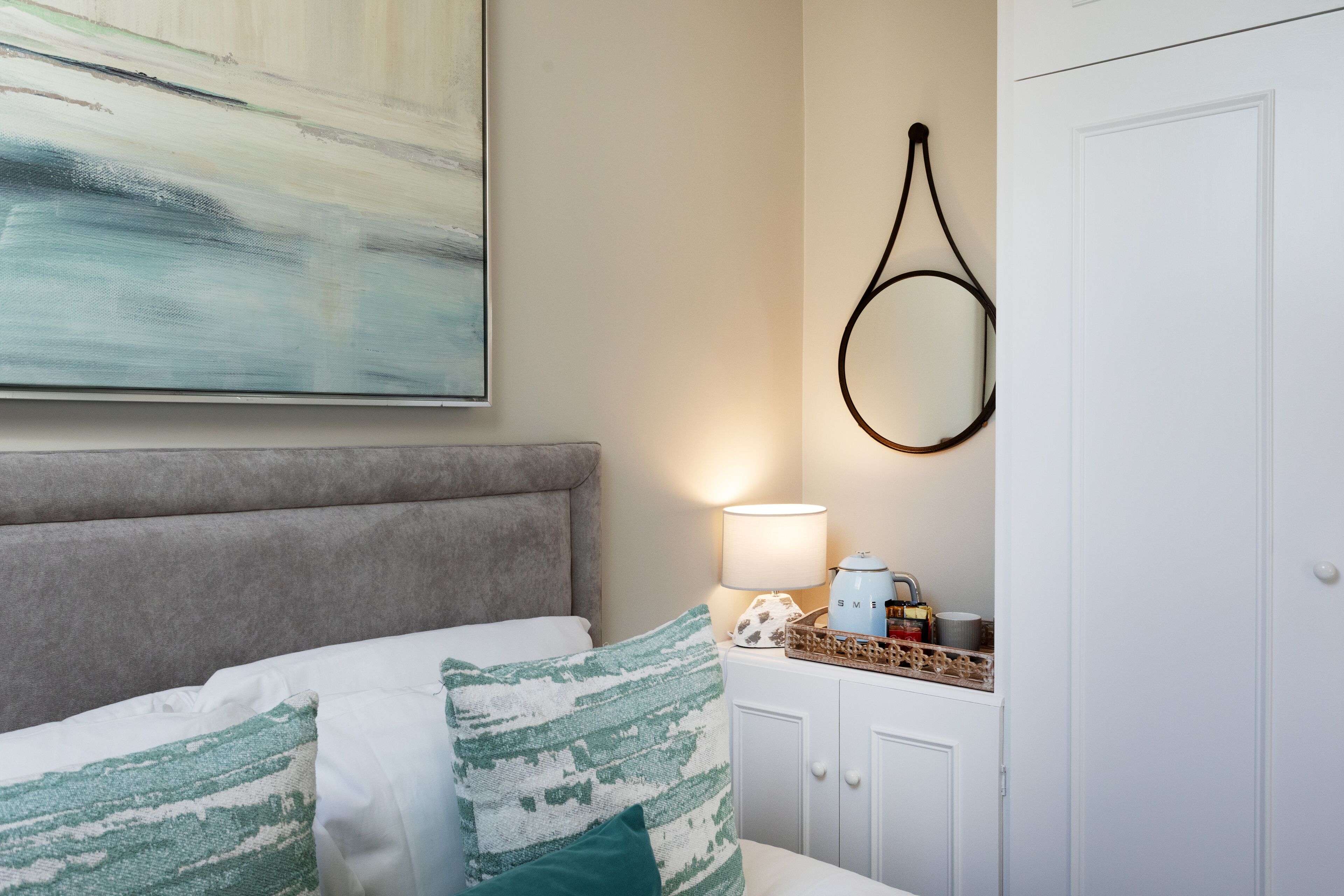 A neatly arranged bedroom with a bed, a bedside lamp, a wall-mounted mirror, and a painting.