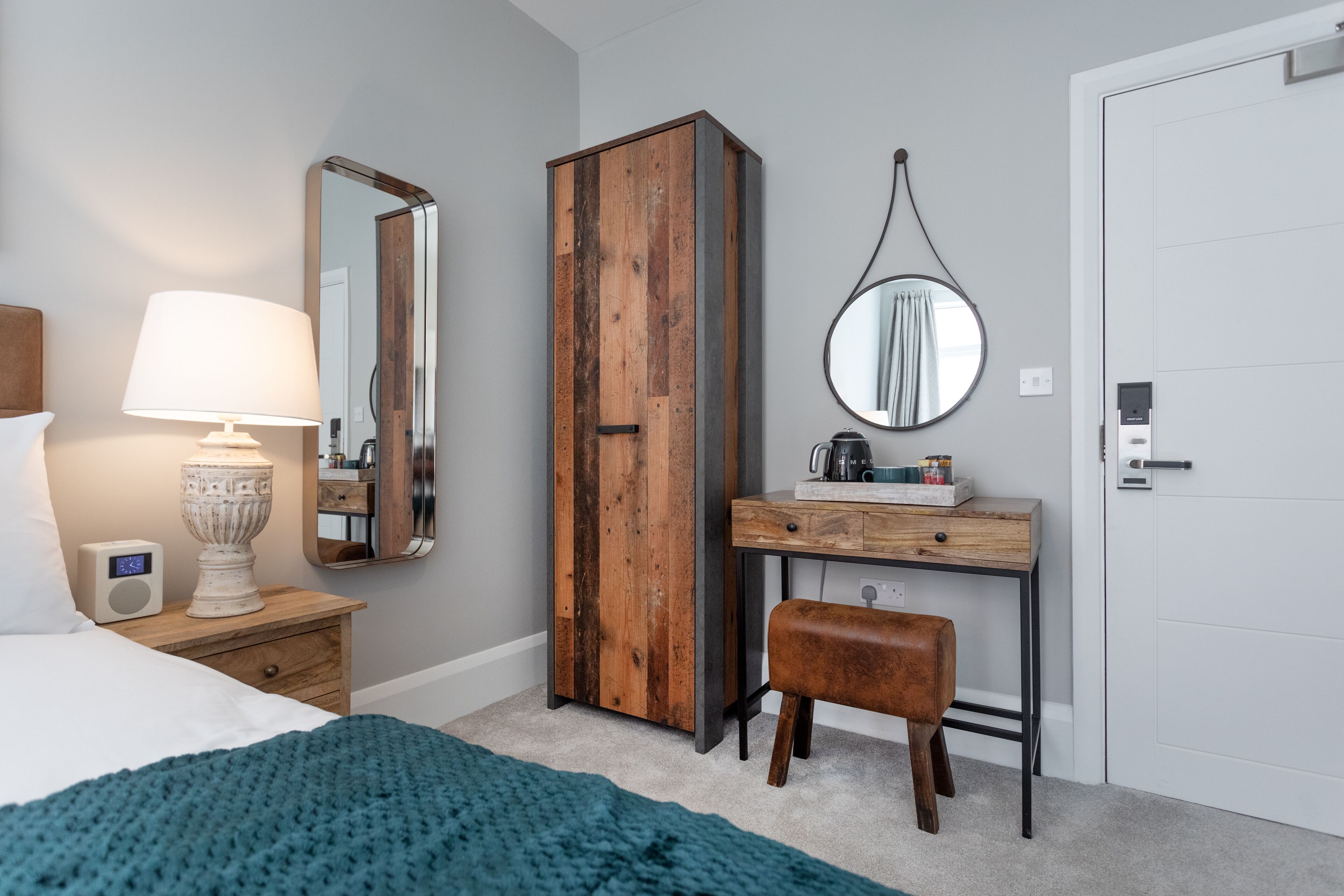 A bedroom with a bed, bedside table with a lamp, a wardrobe, a desk with a mirror, and a door.