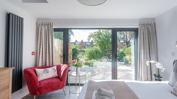 A bedroom with a large bed, a red armchair, a small table with a vase of flowers, and a view of a garden and buildings outside. - No.192 Oxford (Oxford)