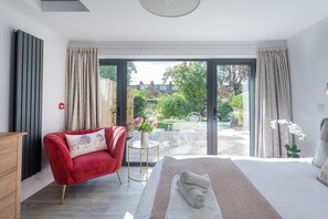 A bedroom with a large bed, a red armchair, a small table with a vase of flowers, and a view of a garden and buildings outside.