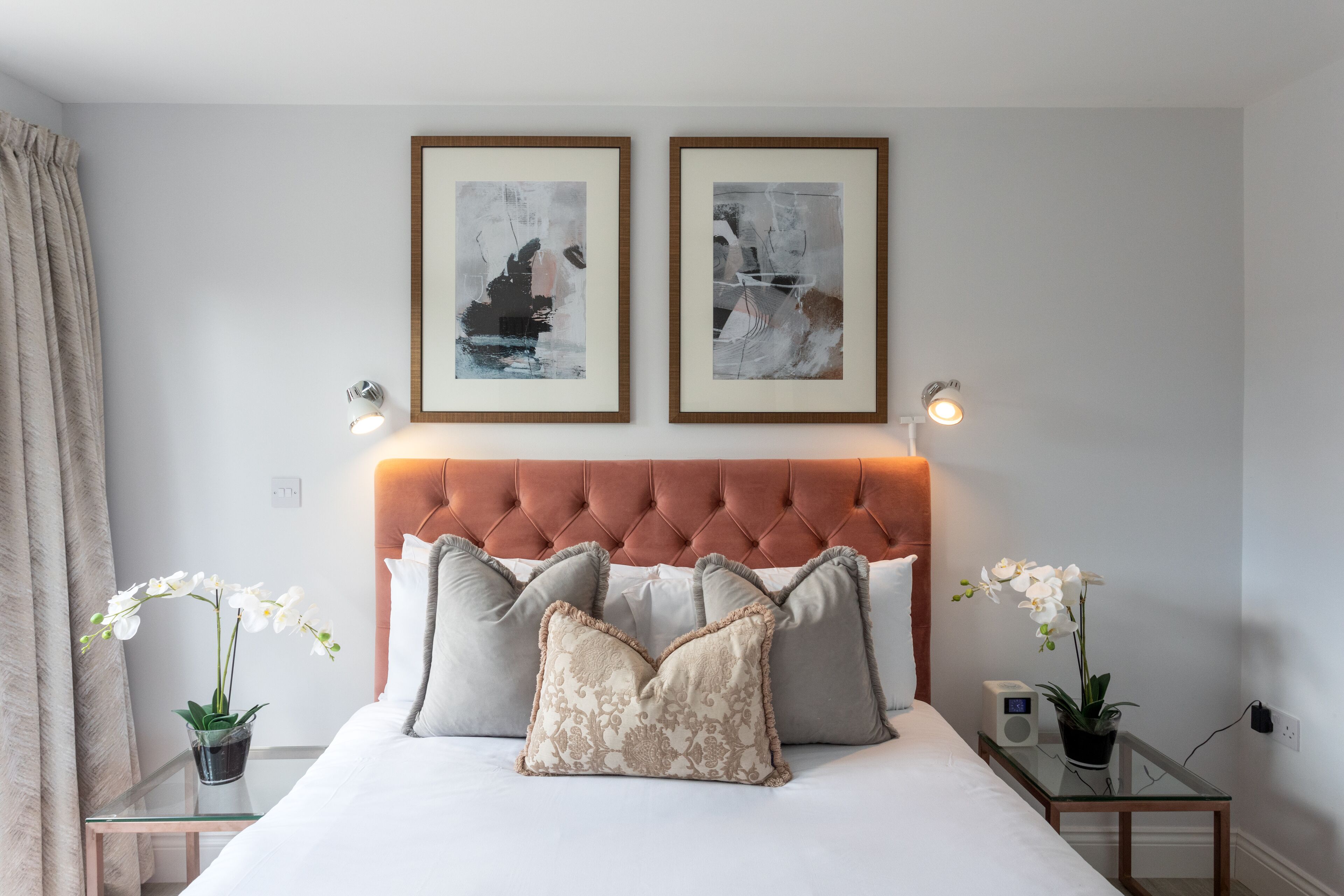A neatly arranged bedroom with a tufted headboard, a glass bedside table, a bed with white bedding, two framed abstract paintings, and two potted orchids.